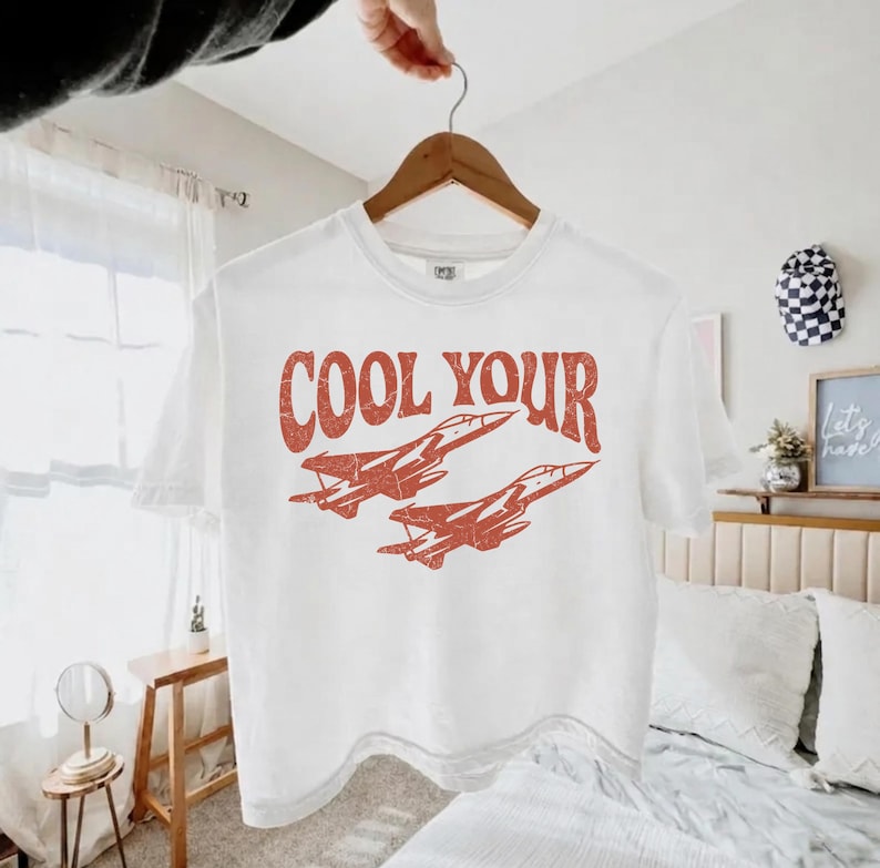 May include: A white short-sleeve t-shirt featuring a distressed red graphic of two fighter jets flying in formation. Above the jets, the text reads COOL YOUR in a bold, retro-style font. The shirt is displayed on a wooden hanger.