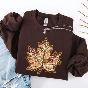 Peut inclure: Un sweat-shirt marron orné d'une grande feuille d'érable détaillée. La feuille est composée de divers motifs à carreaux et floraux aux couleurs automnales, notamment le rouge, l'orange et le vert. Un collier argenté avec un petit pendentif repose sur le sweat-shirt.