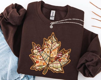 May include: A brown sweatshirt featuring a large, detailed maple leaf design. The leaf is composed of various plaid and floral patterns in fall colors, including red, orange, and green. A silver necklace with a small pendant rests on the sweatshirt.