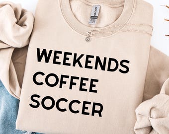 Puede incluir: Una sudadera beige con las palabras "WEEKENDS COFFEE SOCCER" impresas en negro. Un collar plateado con un colgante en forma de sol descansa sobre la sudadera. La sudadera está doblada sobre unos vaqueros azules.