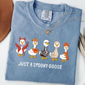 May include: A blue Comfort Colors t-shirt featuring a graphic of five cartoon geese dressed in Halloween costumes. The text "JUST A SPOOKY GOOSE" is printed below the geese. A gold necklace is draped across the shirt.
