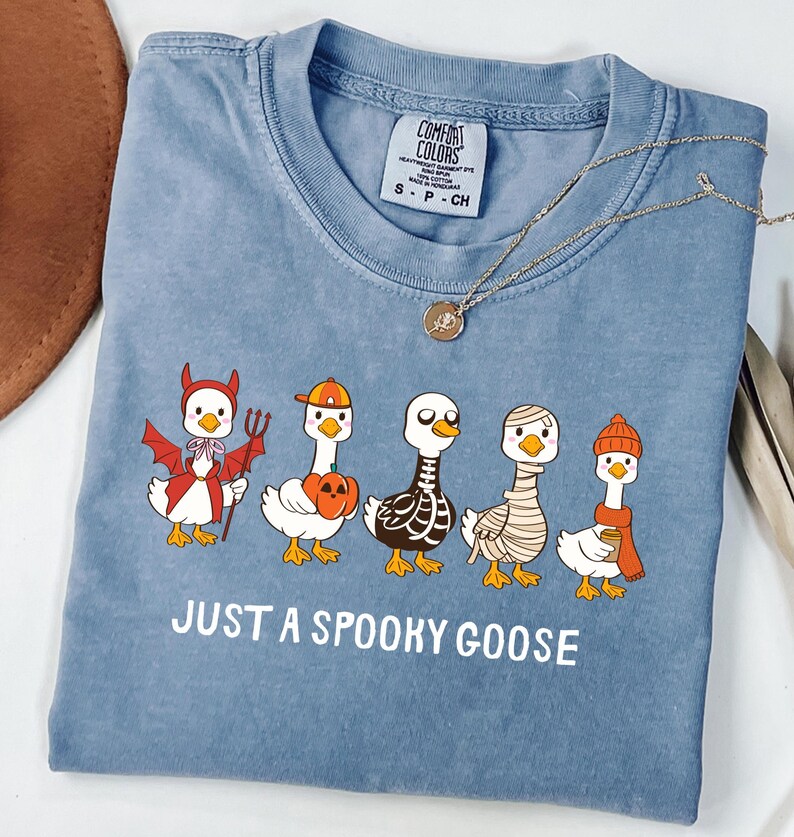 May include: A blue Comfort Colors t-shirt featuring a graphic of five cartoon geese dressed in Halloween costumes. The text "JUST A SPOOKY GOOSE" is printed below the geese. A gold necklace is draped across the shirt.