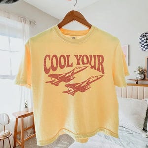 May include: A pale yellow short-sleeve t-shirt featuring a distressed red graphic of two fighter jets flying in formation. The text "COOL YOUR" is printed in a bold, arched, distressed red font above the jet illustration on the front.