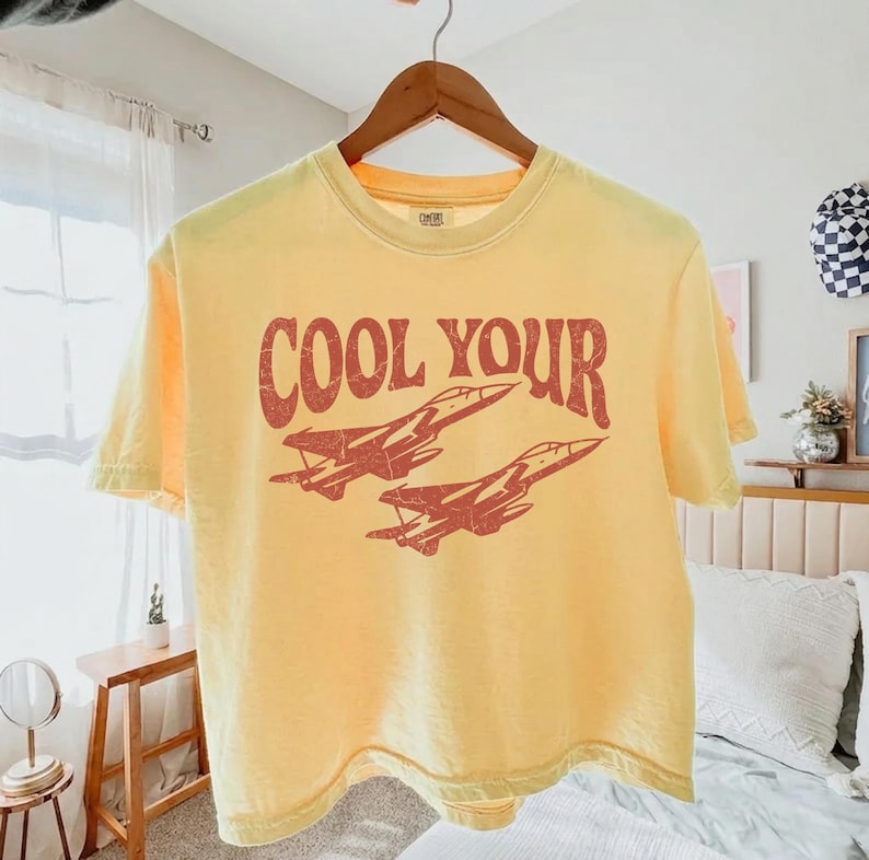 May include: A pale yellow short-sleeve t-shirt featuring a distressed red graphic of two fighter jets flying in formation. The text "COOL YOUR" is printed in a bold, arched, distressed red font above the jet illustration on the front.