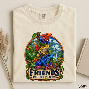 May include: An ivory t-shirt featuring a circular graphic of three colourful frogs perched on a tree branch amidst a tropical landscape. The frogs are red, yellow, and blue. Below the frogs, the text reads "FRIENDS IN HIGH PLACES" in dark blue.