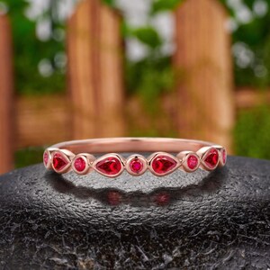 May include: A rose gold band ring featuring an alternating pattern of round and pear-shaped red gemstones set in a bezel style. The ring is positioned horizontally, showcasing the vibrant red stones and the polished metallic finish of the band.