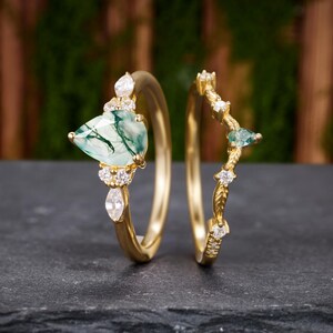 Puede incluir: Un conjunto de anillos de oro con una piedra central de ágata musgosa en forma de pera y piedras blancas en el aro, junto a una alianza de oro curva a juego decorada con pequeñas piedras blancas y una pequeña piedra verde en el centro.