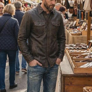 Puede incluir: Chaqueta camisa de cuero marrón con dos bolsillos en el pecho y cierre de botones. La chaqueta se lleva con vaqueros azules. La imagen está tomada al aire libre, con un fondo borroso de personas y puestos de mercado.