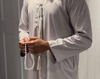May include: A light gray thobe, a traditional Arab garment, with long sleeves and a mandarin collar. The thobe features a tassel detail at the neckline and a pocket on the side. A string of white prayer beads is held in the hands.