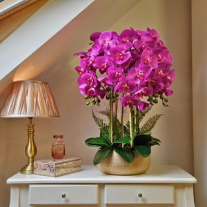May include: A magenta orchid arrangement in a gold-toned textured pot. The artificial flowers are clustered, with green leaves and fern accents. A gold lamp and decorative items sit on a white chest of drawers.