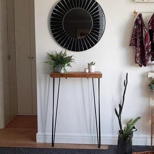 Può includere: Uno specchio rotondo nero con un design a raggiera è appeso sopra una consolle in legno con gambe a forcina nere. Un vaso di felce e piccole piante grasse sono sul tavolo. La stanza ha pareti bianche e un tappeto grigio scuro.