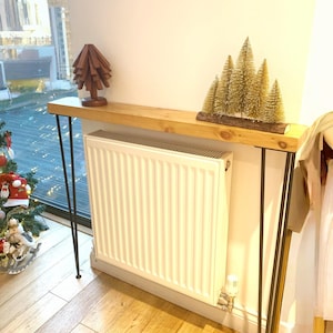 Peut inclure: Une console en bois avec des pieds en épingle noirs. La table est surmontée d'une étagère en bois, affichant un arbre en bois et un groupe de petits arbres de Noël dorés. Un radiateur blanc est en dessous.