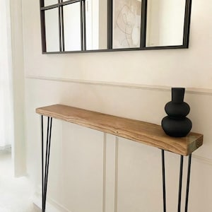 Peut inclure: Une console en bois avec des pieds en épingle noirs. Le plateau est de couleur marron clair. Un vase noir à deux niveaux est posé sur la table. Un miroir noir encadré est suspendu au-dessus de la table.