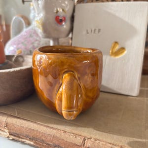 Peut inclure: Une tasse en céramique ambrée et brillante, dotée d'une forme irrégulière façonnée à la main et d'une protubérance sculptée proéminente sur le devant. La surface présente un vernis lisse et réfléchissant avec des variations de tons subtiles.