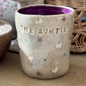 Può includere: Una tazza in ceramica beige maculata con un interno viola vivace. L'esterno presenta piccoli fiori dipinti in bianco, giallo e viola, con il testo THE AUNTIE impresso in lettere maiuscole sulla parte anteriore del contenitore cilindrico.