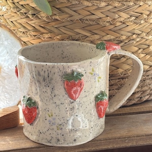 Op de afbeelding: Een gespikkelde keramische mok met een aardbeienontwerp. De mok heeft verhoogde rode aardbeien met groene bladeren. De handgreep heeft ook een aardbeienaccent. De mok is gebroken wit met zwarte spikkels.
