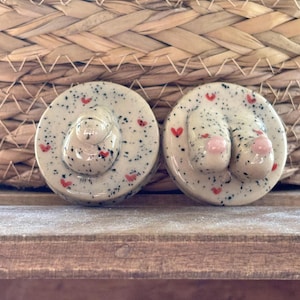 Peut inclure: Deux bouchons de bouteille en céramique. Chaque bouchon est rond, moucheté de points noirs et décoré de petits cœurs rouges. L'un a un motif de chapeau, l'autre un motif de pied avec des orteils roses.