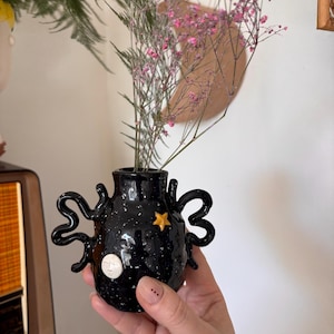 Könnte beinhalten: Eine kleine, schwarze Keramikvase mit gesprenkelter Oberfläche, verziert mit einem gelben Stern und einem weißen Mondgesicht. Die Vase hat einzigartige, geschwungene Griffe und ist mit zarten rosa Blüten gefüllt. Ein skurriles, dekoratives Stück.
