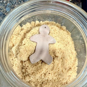 May include: A clear glass jar filled with brown sugar, with a small, light brown gingerbread man-shaped object resting on top of the sugar. The jar is partially visible, showing the rim and the contents within.