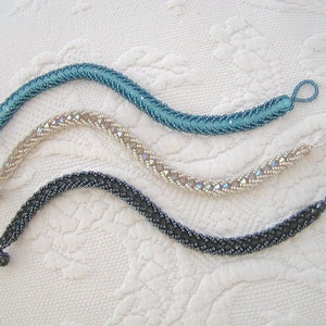 Könnte beinhalten: Drei Perlenarmbänder in verschiedenen Farben: Türkis, Creme und Schwarz. Jedes Armband hat ein einzigartiges Muster und ist aus kleinen Perlen gefertigt.