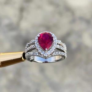 以下が含まれることがあります： 中央に大きな洋梨型の濃い赤色の宝石をあしらったシルバーリング。周囲には小さなクリアストーンのハローが施されています。リングはマルチバンドデザインで、バンドに沿ってクリアストーンがセットされています。