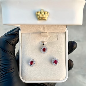 May include: A jewelry set featuring a pendant and two earrings, each with a central red gemstone surrounded by a halo of smaller, clear stones. The set is displayed in a white box with a gold crown emblem.