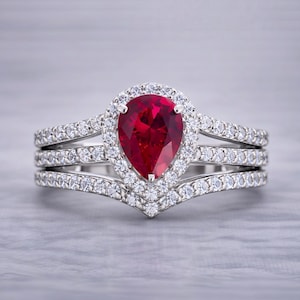 Puede incluir: Un juego de anillos de tres piezas con una piedra preciosa roja oscura en forma de pera, rodeada por un halo de piedras claras más pequeñas. Las bandas están adornadas con piedras claras adicionales, creando un efecto brillante. El anillo está engarzado en una banda plateada.