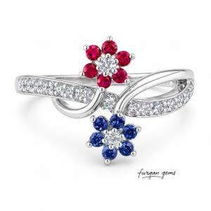 May include: A silver ring featuring two flower designs. One flower is composed of red gemstones, the other of blue gemstones, each with a central white gemstone. The ring band is adorned with small white gemstones.