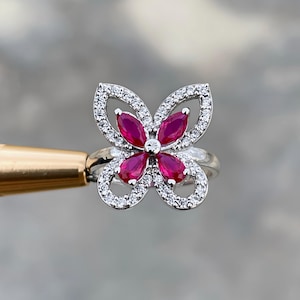 May include: A silver ring featuring a butterfly design. The butterfly's wings are outlined with small, clear gemstones, and the center holds five marquise-cut, deep red gemstones. The ring is set against a blurred background.