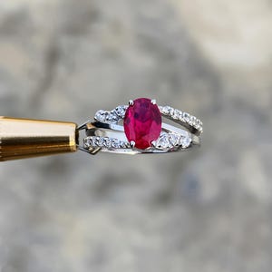 Pode incluir: Um anel de prata com um grande rubi oval como pedra central. O anel é adornado com pedras preciosas redondas e transparentes menores, incrustadas ao longo da banda, criando um efeito brilhante. O rubi é de cor vermelha vibrante.