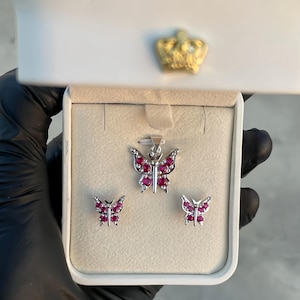 May include: A silver butterfly pendant necklace and matching earrings set, displayed in a white jewelry box. The butterfly designs are adorned with red gemstones. The box has a gold crown emblem.