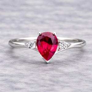 Puede incluir: Un anillo de plata con una piedra preciosa roja vibrante en forma de pera como pieza central, flanqueada por dos piedras blancas redondas más pequeñas. La banda del anillo es delgada y pulida, creando un diseño clásico y elegante.