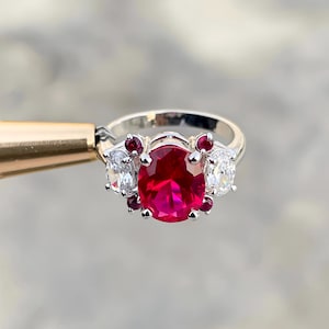 Könnte beinhalten: Ein silberner Ring mit einem großen, oval geschliffenen, leuchtend roten Edelstein im Mittelpunkt. Flankiert wird der zentrale Stein von zwei weißen, oval geschliffenen Edelsteinen, mit kleineren roten Steinen als Akzent. Der Ring wird von einem goldfarbenen Werkzeug gehalten.