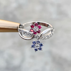 May include: A silver ring featuring two flower designs. The top flower is composed of red gemstones, while the bottom flower is blue. The ring band is adorned with small, clear gemstones, adding sparkle to the floral design.