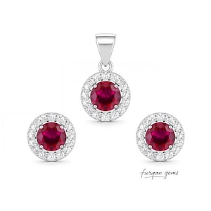 May include: A jewelry set featuring a pendant and two stud earrings. Each piece has a large, round, deep red gemstone surrounded by a halo of smaller, clear gemstones. The set is set in silver-colored metal. The brand name "furqan gems" is written in cursive.