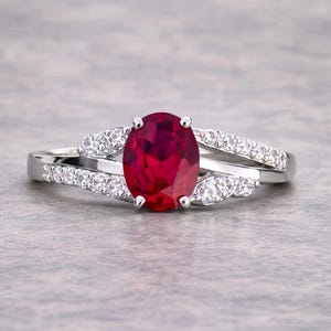 Puede incluir: Anillo de plata con un gran rubí ovalado en el centro. La banda se divide, cada lado adornado con pequeñas piedras blancas. El rubí es de un rojo intenso, y el anillo se muestra sobre un fondo neutro.