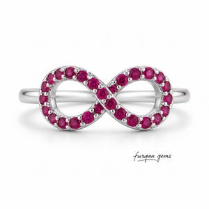 May include: A silver-colored ring featuring an infinity symbol design. The infinity symbol is encrusted with numerous small, round, bright pink gemstones. The ring has a smooth, polished band and the text "furgan gems" is visible.