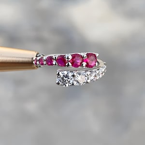 Puede incluir: Un anillo plateado con dos filas de piedras preciosas. Una fila presenta piedras rojas vibrantes, la otra piedras claras y brillantes. El anillo está sostenido por una herramienta dorada sobre un fondo borroso.