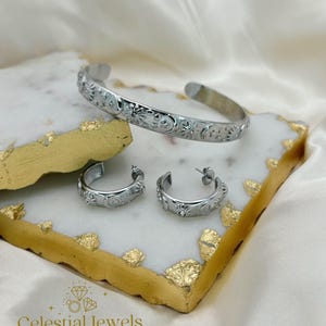 May include: A silver jewellery set comprising a cuff bracelet and two hoop earrings. The bracelet and earrings are decorated with celestial designs of moons, stars, and suns. The jewellery sits on a white and gold decorative tray, with the brand name "Celestial Jewels" visible.
