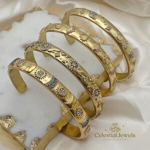 May include: Four gold bangles with celestial designs, including suns, moons, and stars. The bangles are decorated with small, sparkling gemstones in various colours. The jewellery is presented on a white surface.