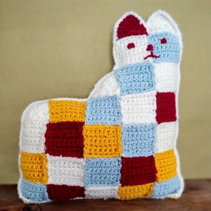 May include: A crocheted cat pillow with a patchwork design in white, red, yellow, and blue.