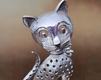 May include: A silver figurine of a spotted cat with large eyes. The cat is sitting on a small base and has a playful expression.