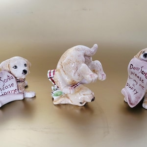 May include: Three ceramic puppy figurines in various poses. Each puppy is cream-colored with brown accents and a red and white striped collar. One puppy holds a scroll that reads "Dear Santa please bring me puppy".