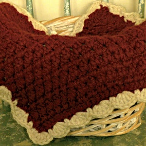 May include: A handmade burgundy crochet blanket with a cream-colored scalloped edge rests in a wicker basket. The blanket's texture is visible, showcasing the intricate stitch work. The basket sits on a distressed green chair, creating a rustic aesthetic.