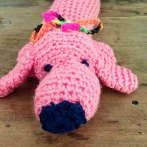 May include: A handmade pink crocheted dog toy with dark blue accents on the nose and eyes. The dog has floppy ears and a colorful collar. The toy is made of soft yarn and is suitable for children.