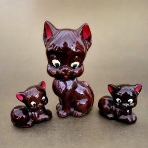 May include: A set of three brown ceramic cat figurines. The largest cat is sitting upright with its ears perked up. The two smaller cats are lying down. All three cats have red painted ears and green eyes.