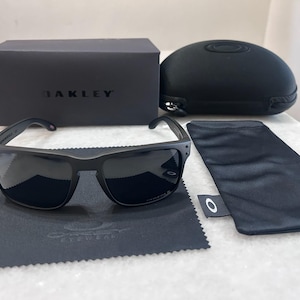 May include: Black Oakley sunglasses with a matching case, box, and cleaning cloth. The sunglasses have dark lenses and a matte black frame. The box and case are also black, with the Oakley logo visible. A black microfiber cloth is included.