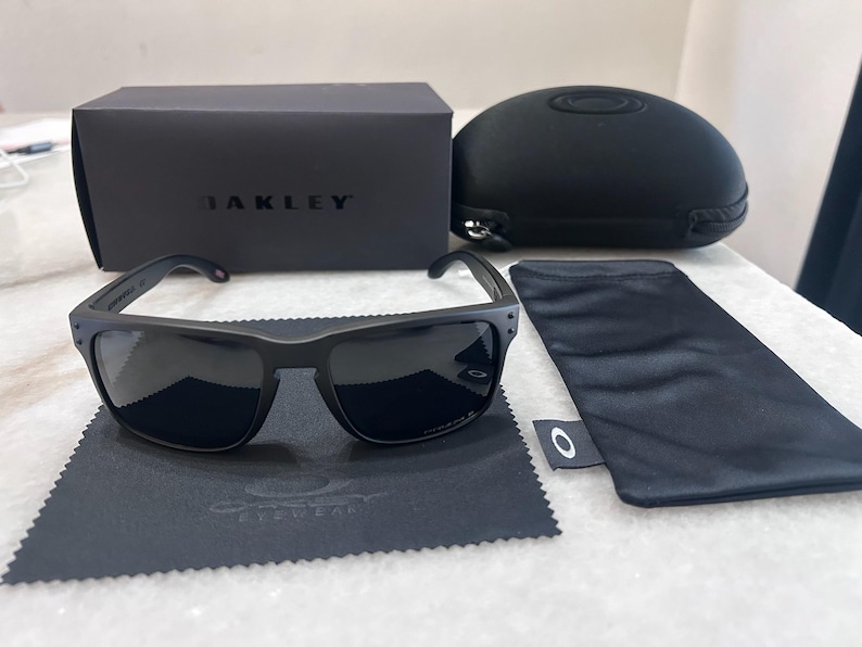 May include: Black Oakley sunglasses with a matching case, box, and cleaning cloth. The sunglasses have dark lenses and a matte black frame. The box and case are also black, with the Oakley logo visible. A black microfiber cloth is included.