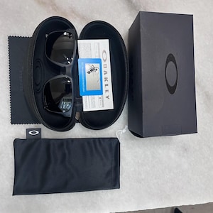 May include: Black Oakley sunglasses in a black case, with a black microfiber cloth, a black pouch, and a black box. The sunglasses are in an open, black, zippered case. The box has the Oakley logo.