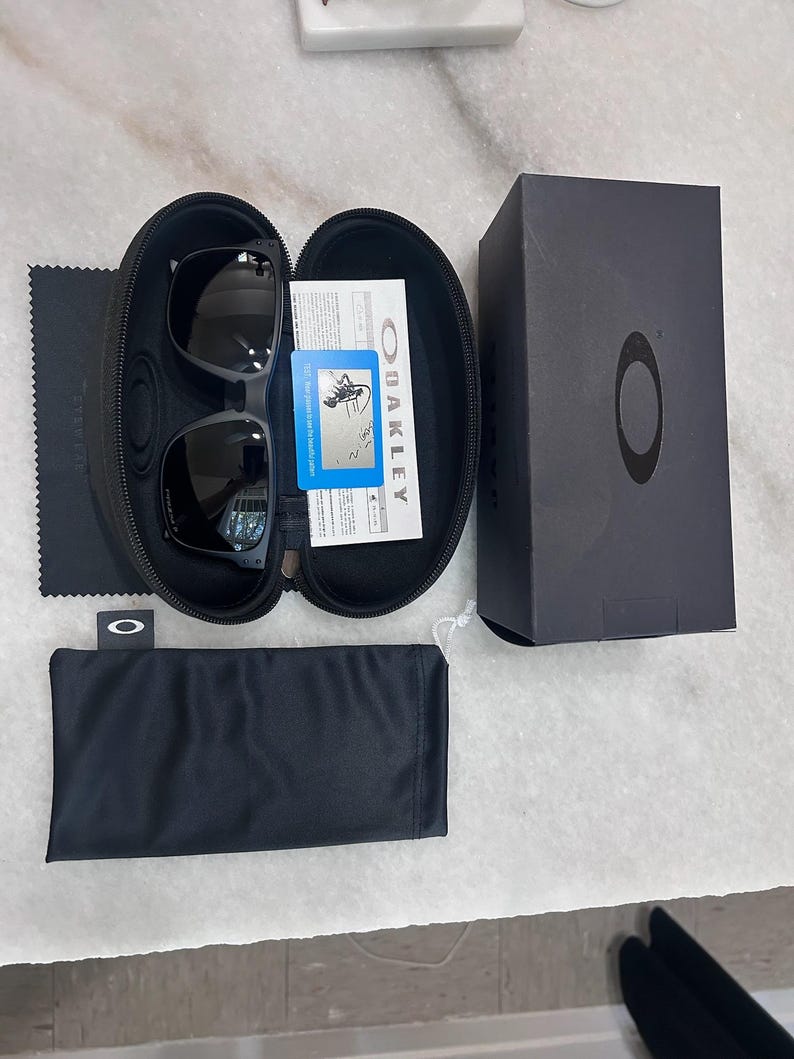May include: Black Oakley sunglasses in a black case, with a black microfiber cloth, a black pouch, and a black box. The sunglasses are in an open, black, zippered case. The box has the Oakley logo.